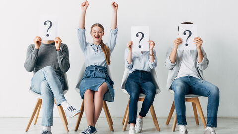 Vier Menschen sitzen auf Stühlen nebeneinander, drei halten eine Karte mit Fragezeichen vor ihr Gesicht. Die vierte Person reißt  die Arme in die Höhe und jubelt