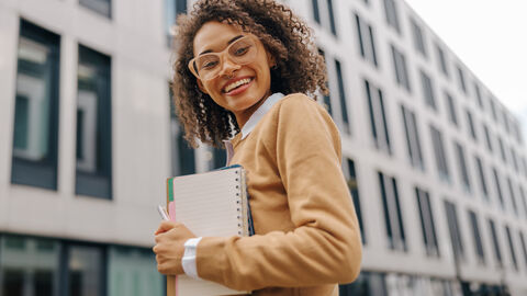 Das HMWK bildet auch aus! Eine Frau steht mit Notizblock lächelnd vor einem modernen Gebäude.