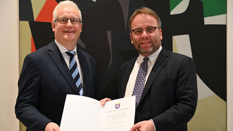 Minister Gremmels steht mit dem neuen Leiter des Hessischen Landesarchivs vor der Kamera und zeigt die Ernennungsurkunde.