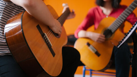 Ein Kind spielt Gitarre, im Hintergrund sitzt eine Erwachsene mit Gitarre und leitet das Kind an