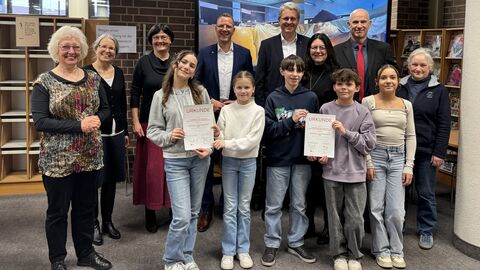 Die Max-Planck-Schule und die Stadtbücherei Rüsselsheim erhalten den Hessischen Leseförderpreis 2024 und 3.000 Euro Preisgeld für ihr Projekt „Jahrmarkt der Bücher“.  Staatssekretär Christoph Degen steht mit den Gewinnerinnen und Gewinnern des Hessischen Leseförderpreises in der Rüsselsheimer Stadtbücherei.