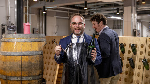 Minister Gremmels hält eine Flasche Wein und steht neben einem Fass