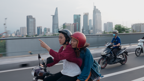 Szene aus "Saigon Kiss": Ein Paar auf einem Motorroller vor der Skyline Saigons